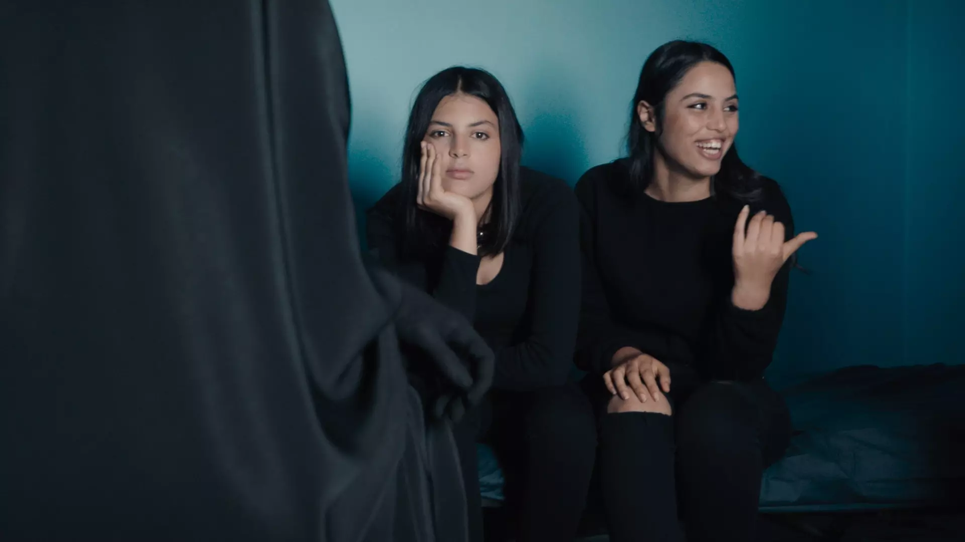 To unge tunisiske kvinner sitter på siden av hverandre på en seng eller sofa. Begge er kledd i sort. 