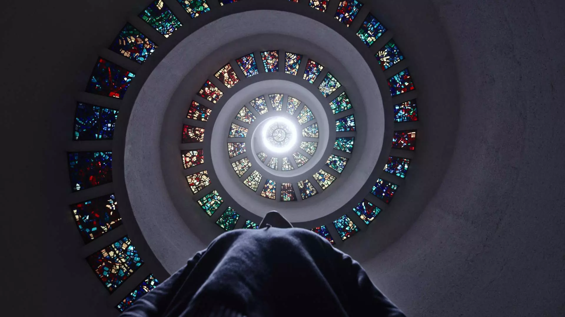 Halvtotal i froskeperspektiv. En person ser oppover i det som ser ut til å være i et religiøst rom. Det ser ut som en spiral av glassmalerier opp mot et vindu i toppen av et tårn, innenifra. Foto.