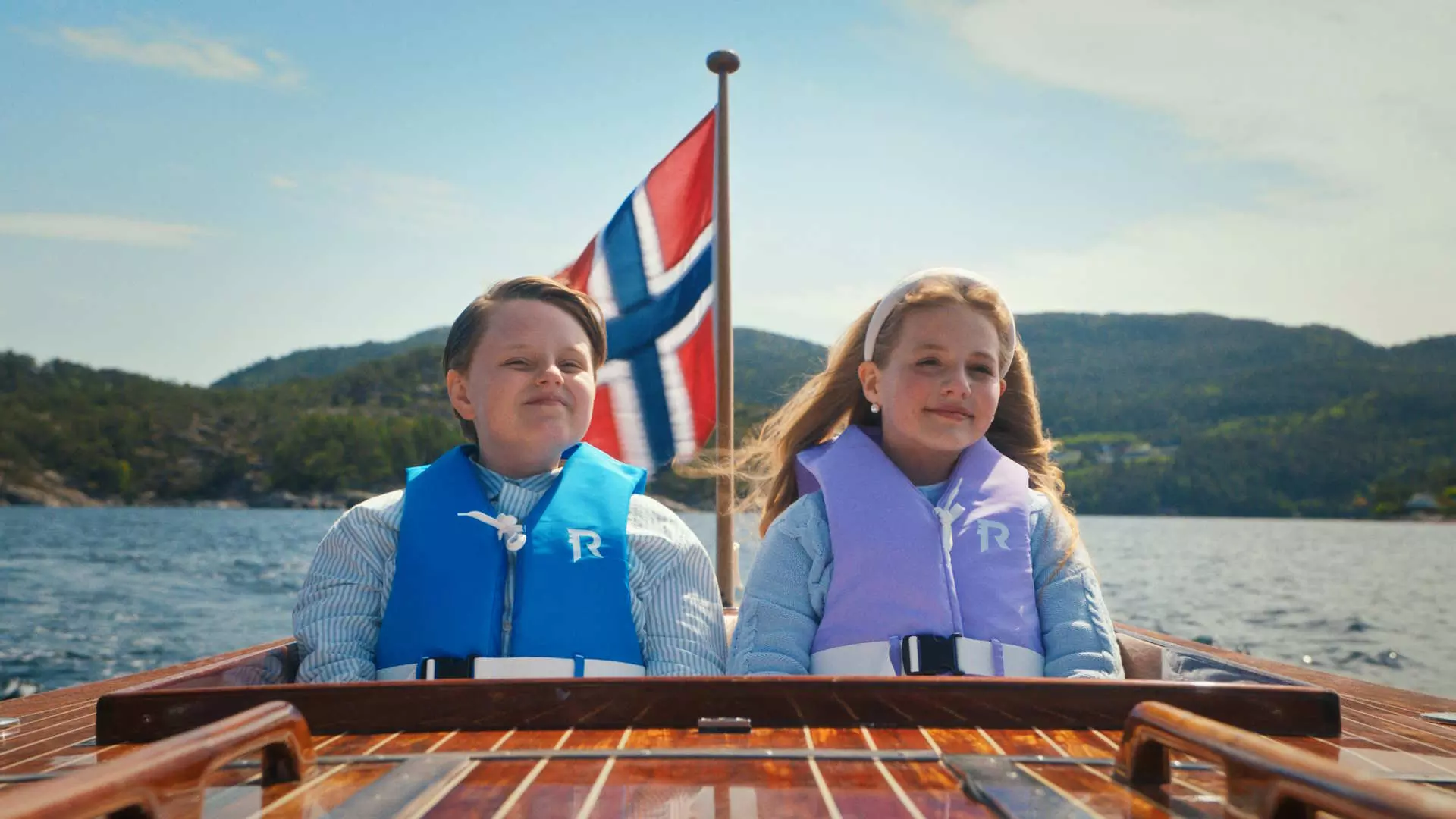 To barn sitter bak i en sjekte. De er i kledd klær og flytevest i pastellfarger. De smiler falskt. Bak dem ser vi et norskt flagg. Det er blå himmel og fjell i bakgrunnen.