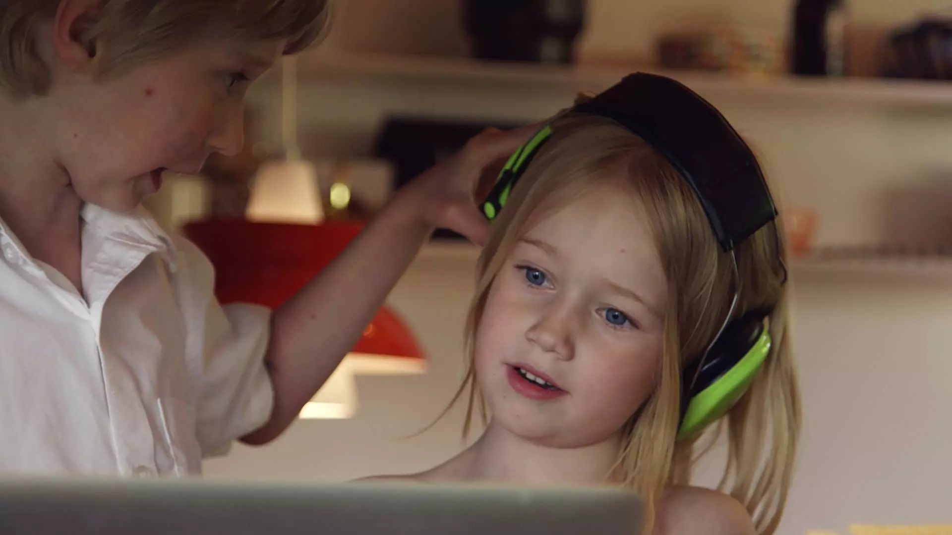 To barn som sitter forran det som kan være en laptop. Jenta har på seg svarte og grønne hodetelefoner. Gutten holder i hodetelefonene hennes. Foto.