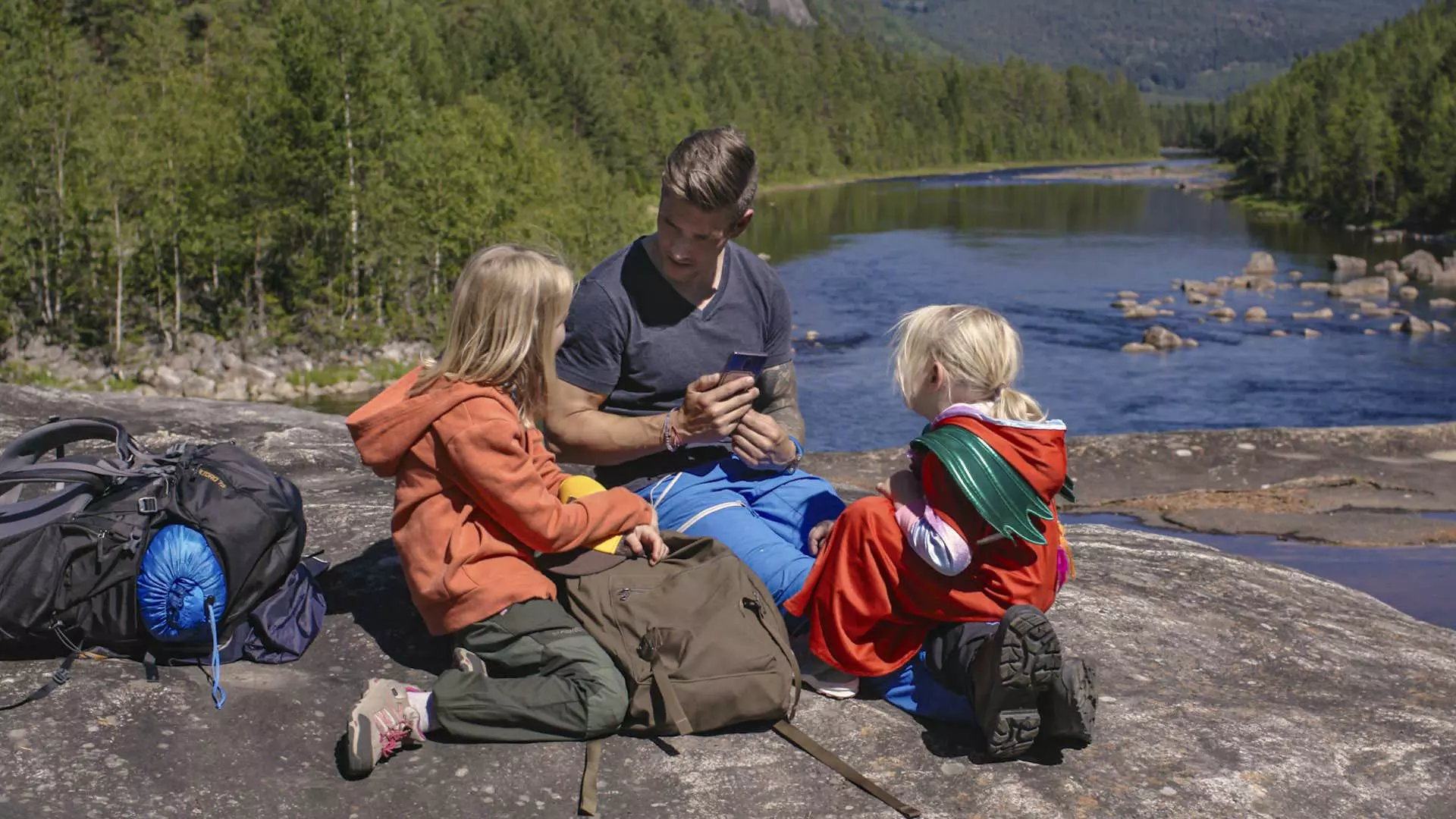 En mann og to barn på 8 og 5 år. De sitter på en stein i skogen. Bak dem er et vann. De har tursekker med seg. 