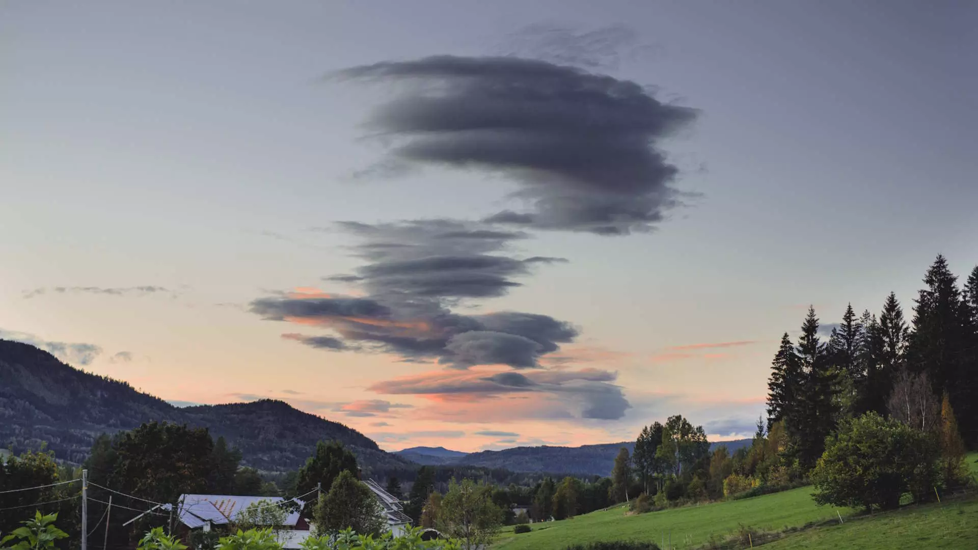 En vakker solnedgang over et landlig landskap med åser, trær og hus. Bildet gir en fredelig og drømmende stemning.