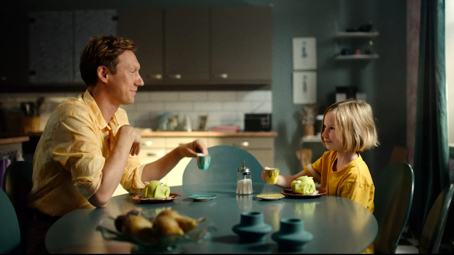En mann og en jente sitter ved frokostbordet med kjøkkenet i bakgrunnen. Begge har på seg gult. Det ser ut som de har det gøy. 