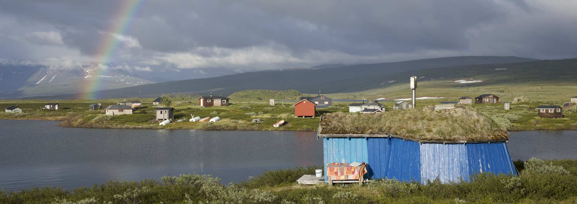 Stillbilde fra "Samiske fortellinger om rett og urett". Nydelig landskap i nord med regnbue, sjø og små hus. Foto.