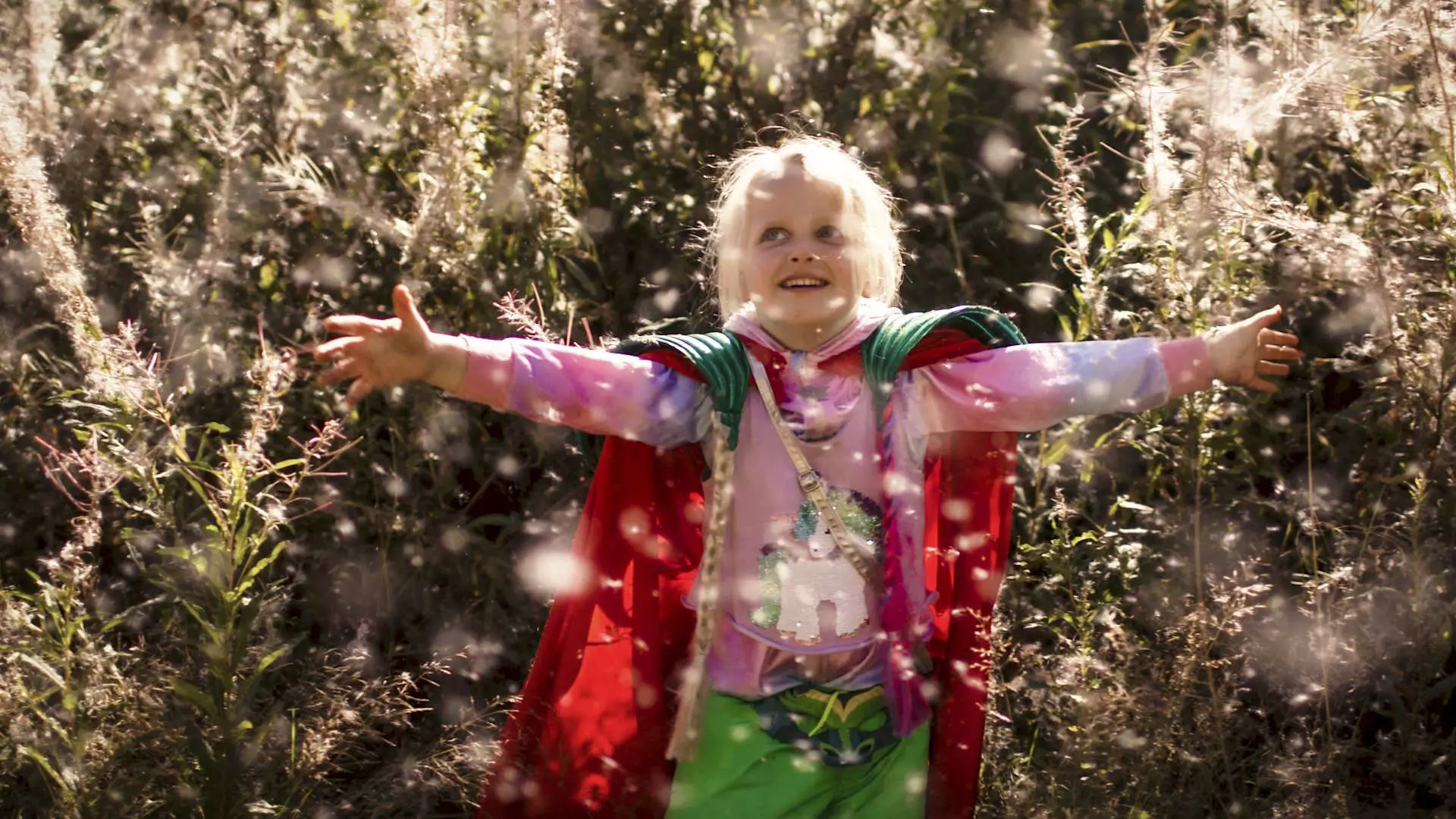 En 5 år gammel jente i skogen. Sterkt spisslys fra solen. Hun står i en eng av strå som flåser små korn i luften. Bildet ser magisk ut. Jenta står med armene rett ut og ser lykkelig ut.