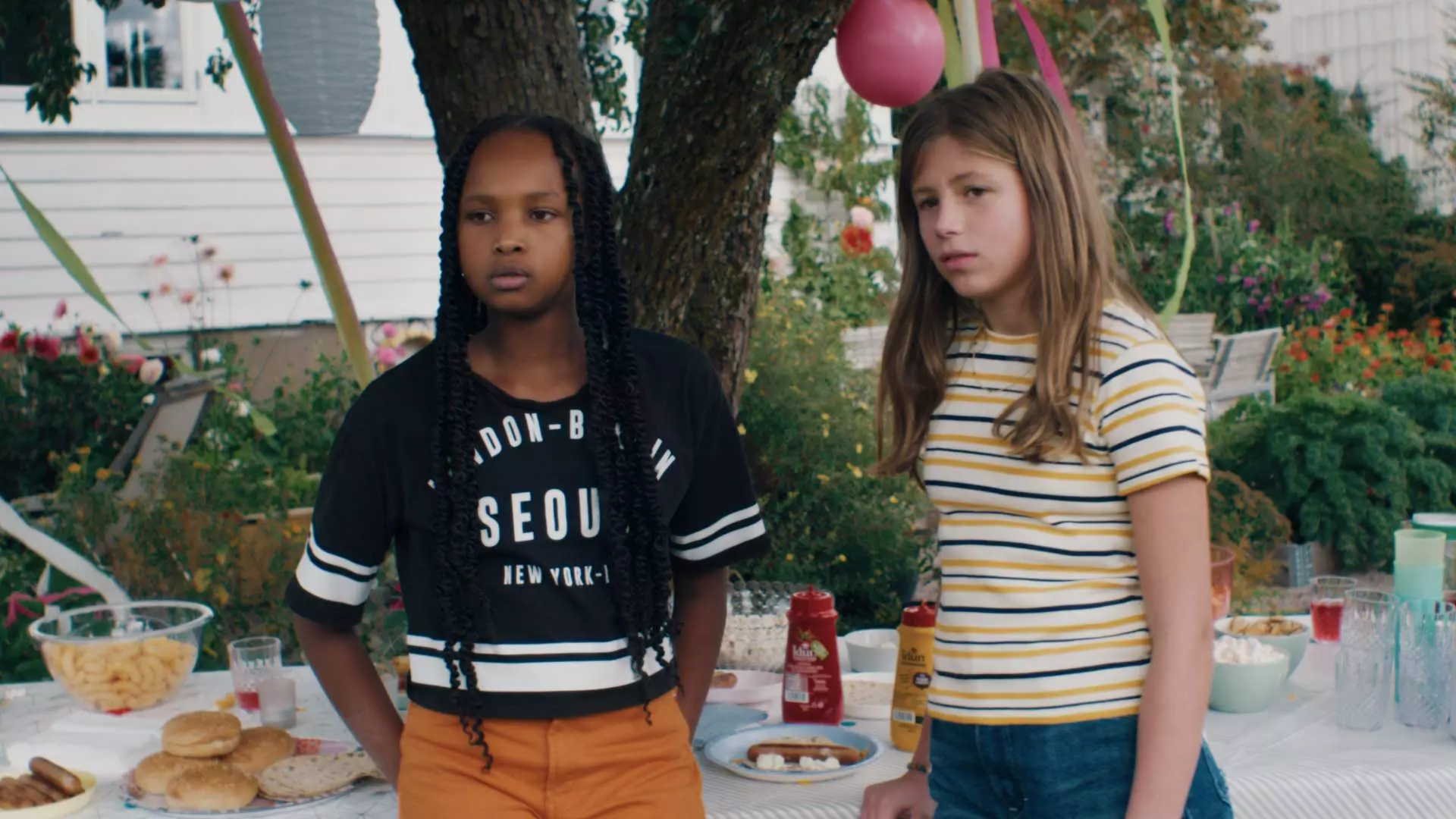 To unge jenter på hagefest. De er omkring 11 år. De står forran et bord med hamburgerbrød, ketchup og annet tilbehør. Det henger ballonger i treet og hagen blomstrer. Jentene har t-skjorte på seg. Det virker som at det er sommer. De ser alvorlige ut.