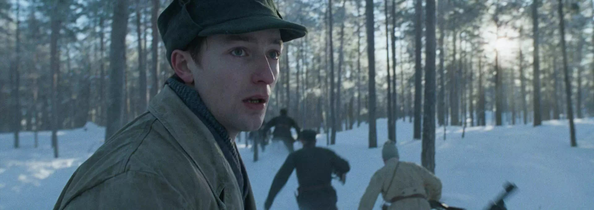 Stillbilde fra filmen "Nr. 24". Snødekket skog. En ung mann ikledd militærklær fra andre verdenskrig snur seg forskrekket over skulderen. I bakgrunnen ser vi flere menn som løper fra noe.