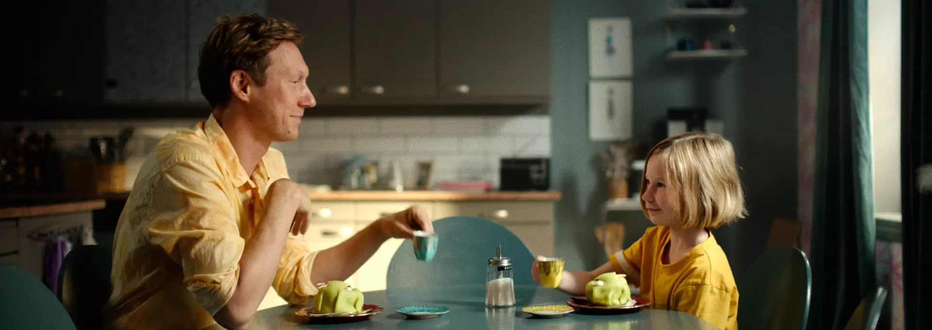 En mann og en jente sitter ved frokostbordet med kjøkkenet  bakgrunnen. Begge har på seg gult. Det ser ut som de har det gøy. 