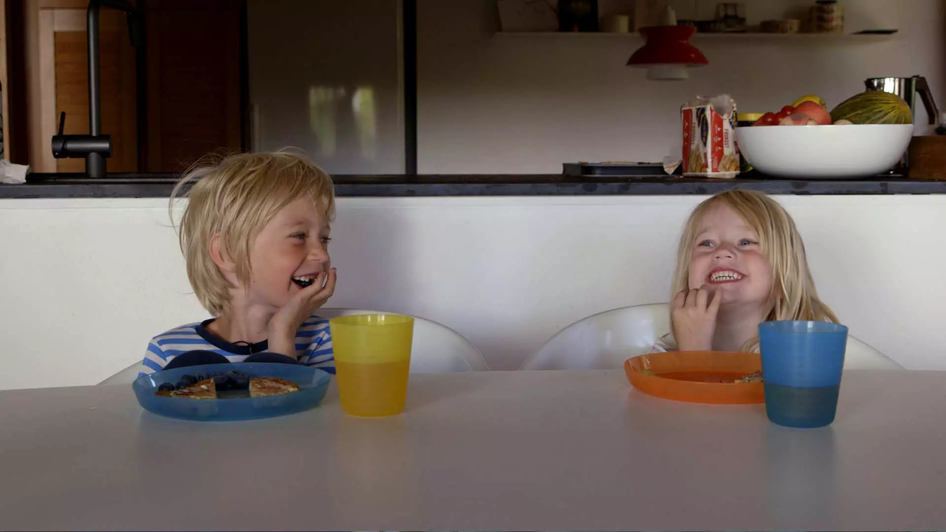 To barn sitter ved frokostbordet. De ler og koser seg. Forran dem på bordet har de hver sin plastkopp og plastikktallerken. Foto