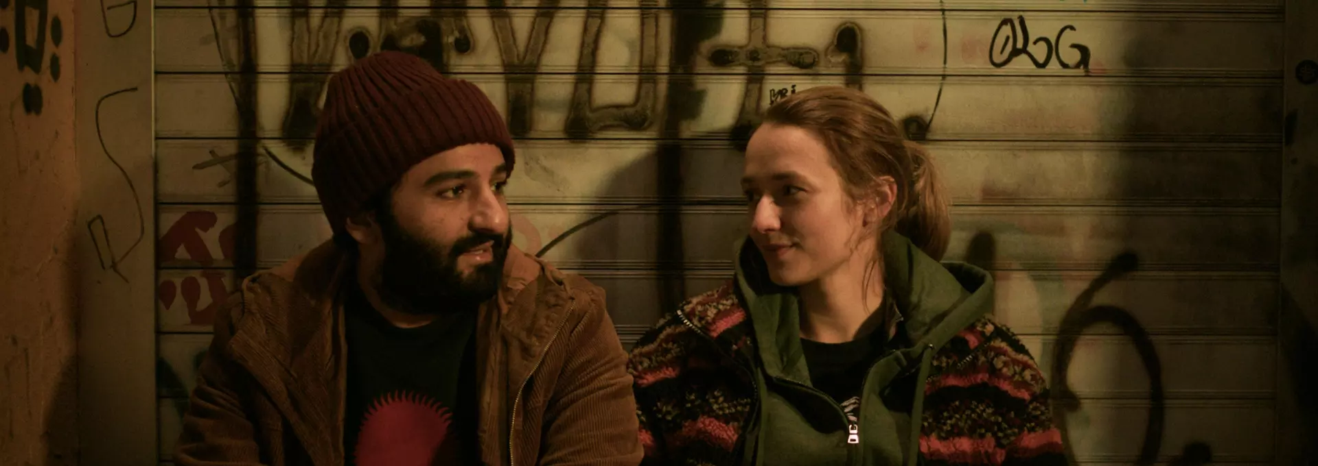 En mann og en kvinne i 20-årene sitter foran en vegg med tagging og prater. 