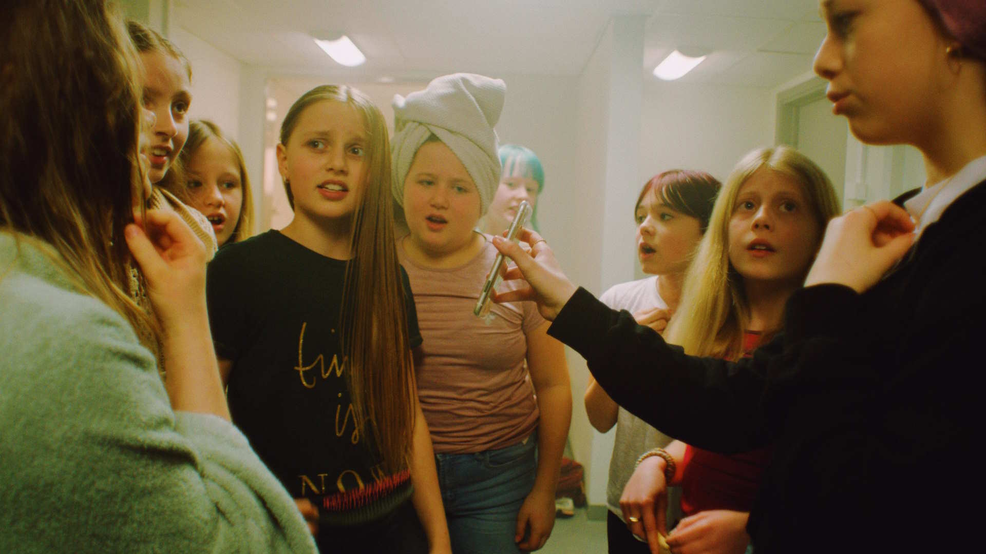 Stillbilde fra "Alle vet". Unge jenter som står samlet rundt en mobiltelefon i en garderobe. Foto.