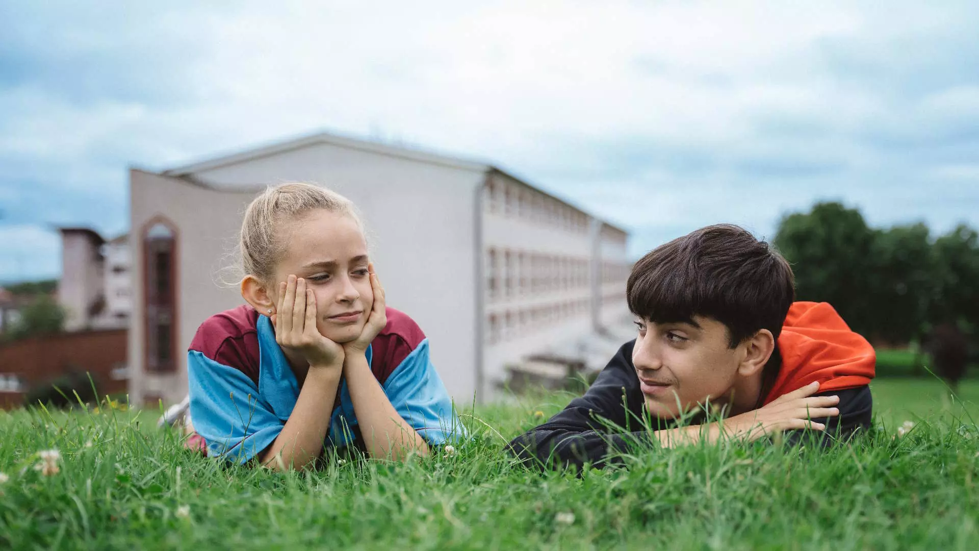 Halvnært. En ung jente og en ung gutt ligger på magen og slapper av på en grønn gressplen. De ser på hverandre. I bakgrunnen kan vi skimte et leilighetskompleks. Det er blå himmel med lette skyer.