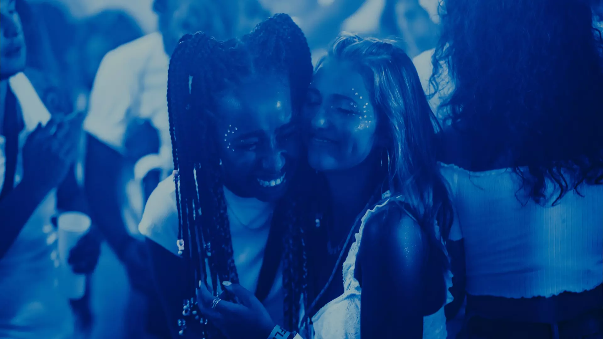 To unge kvinner står på et utested og smiler mens de klemmer hverandre. Det ser ut som det er mørkt og at de er på en klubb sent på kvelden. 