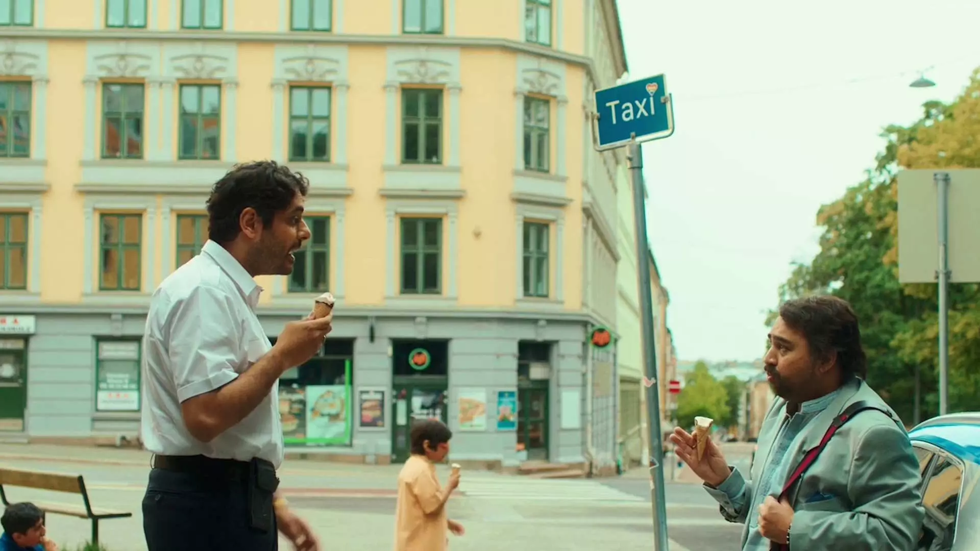 To menn står ved en taxiholdeplass i en bygate. De spiser iskrem om ser på hverandre. I bakgrunnen kan man se en gul bygning og en person som går forbi. Det er dagslys og gaten virker rolig med få mennesker og biler.