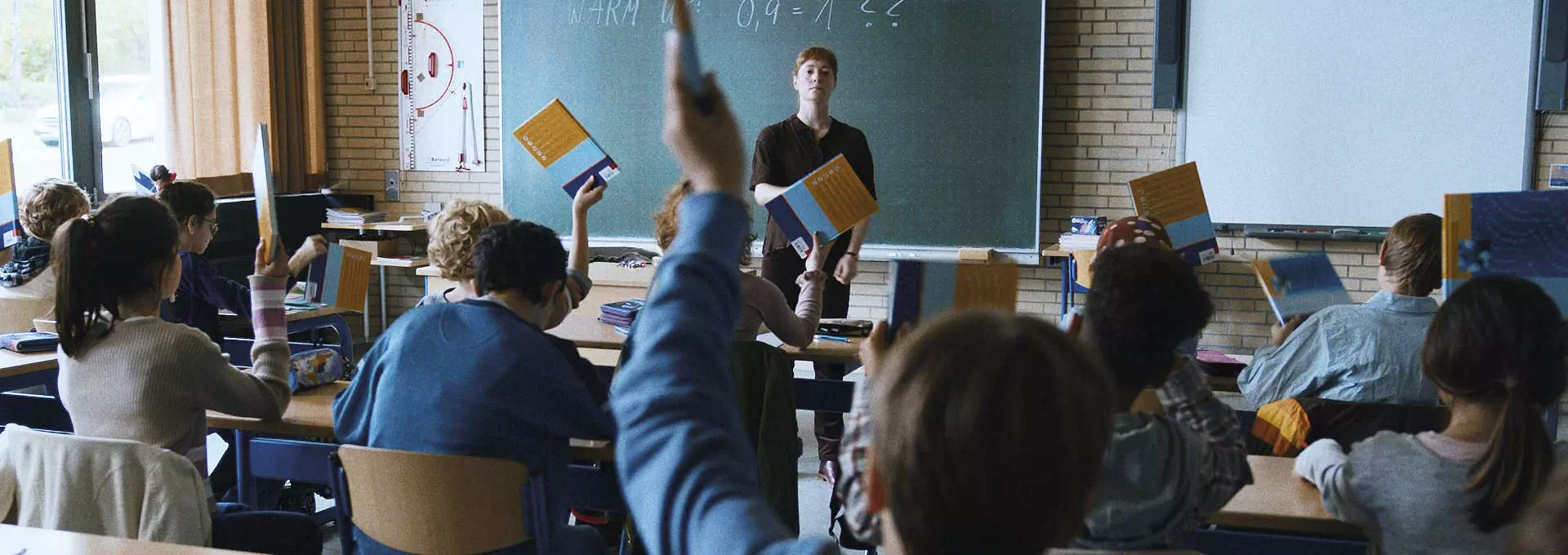 Bildet viser en klasseromssituasjon hvor en lærer står foran klassen mens elevene sitter ved sine pulter. Noen av elevene har reist hånden og holder opp en bok. Klasserommet ser ut til å være godt opplyst med naturlig lys, og det er en tavle i bakgrunnen med skrift. Læreren ser ut til å være oppmerksom på elevene. Atmosfæren ser ut til å være rolig og organisert.