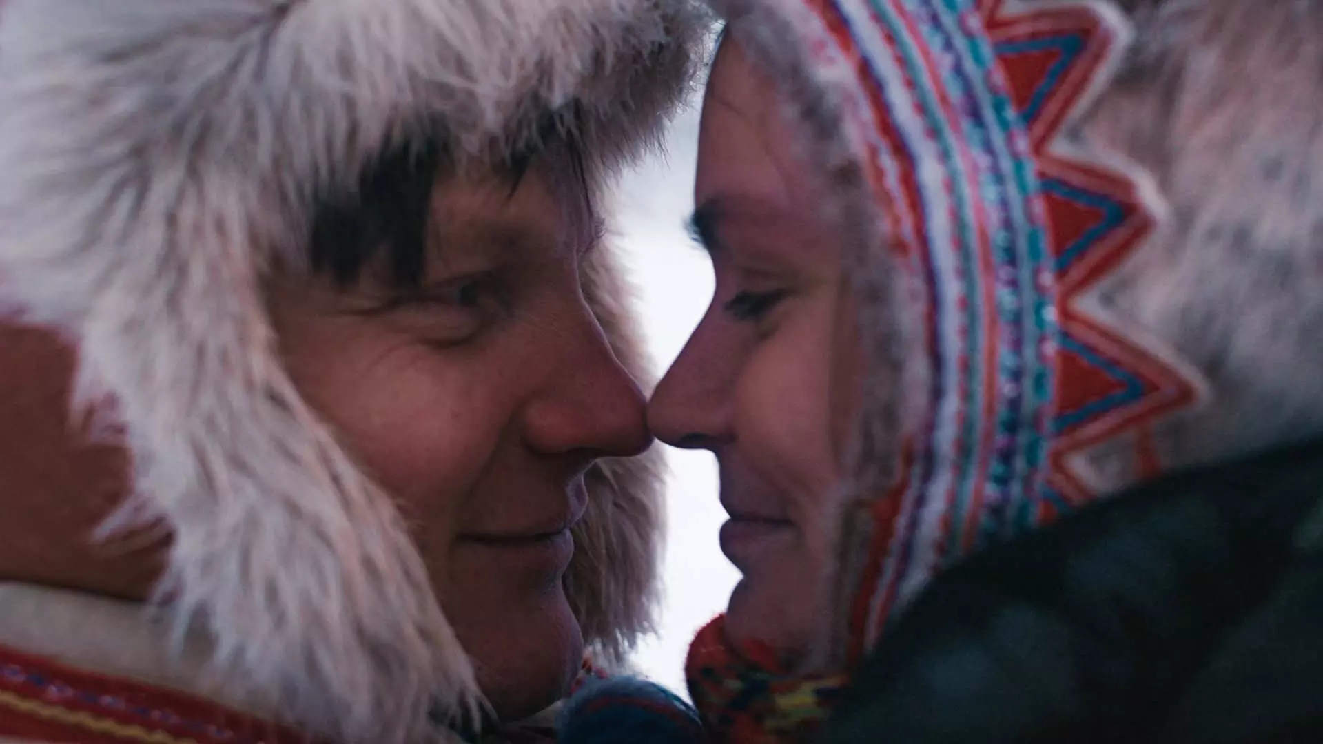 Nærbilde. En ung samisk mann og samisk kvinne i profil. De har nese mot nese og smiler, de ser forelsket ut. De er ikledd tradisjonelle samiske luer. Foto.