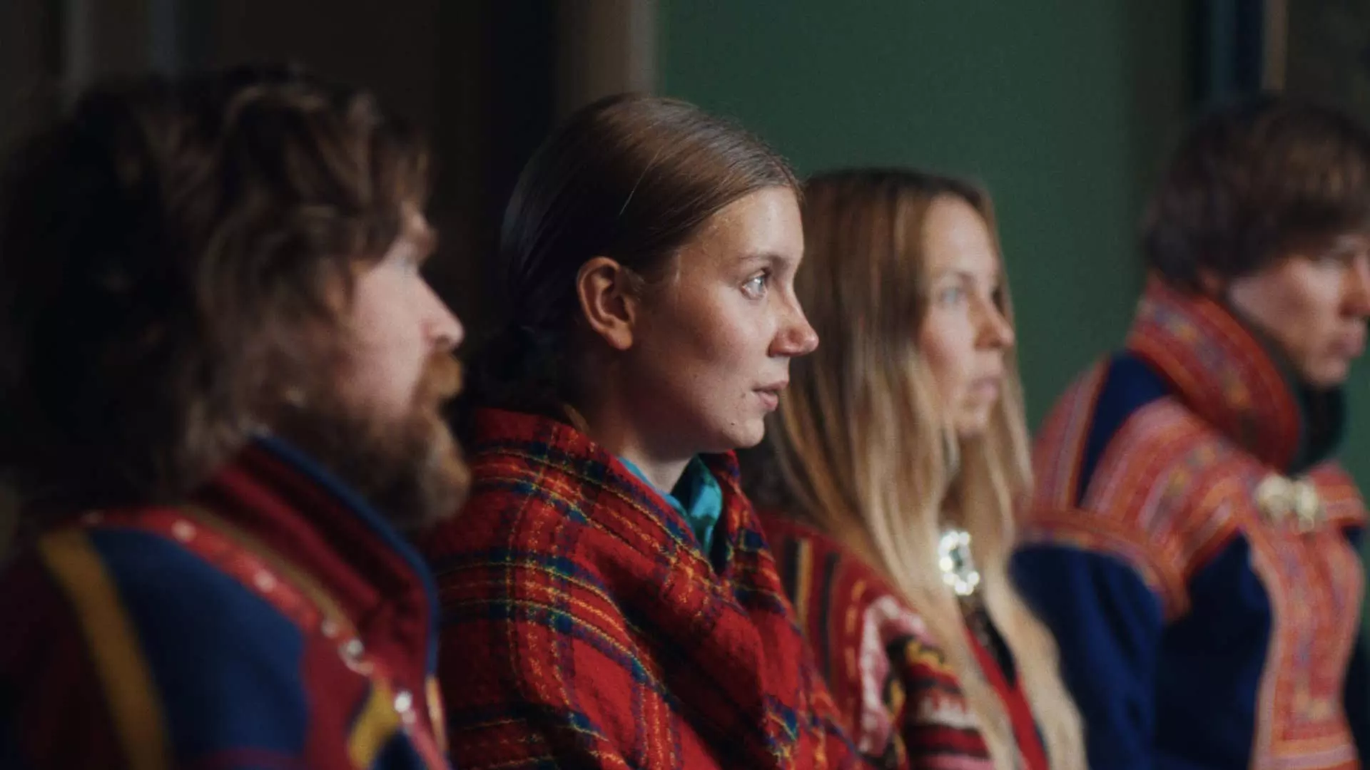 Ung kvinne i profil. Hun har et rødt rutete sjal rundt seg. Det ser ut som et sjal som tilhører en samisk kofte. Hun ser alvorlig ut. Rundt henne sitter flere mennesker som også har på seg samiske kofter. De befinner seg i et grønt rom.