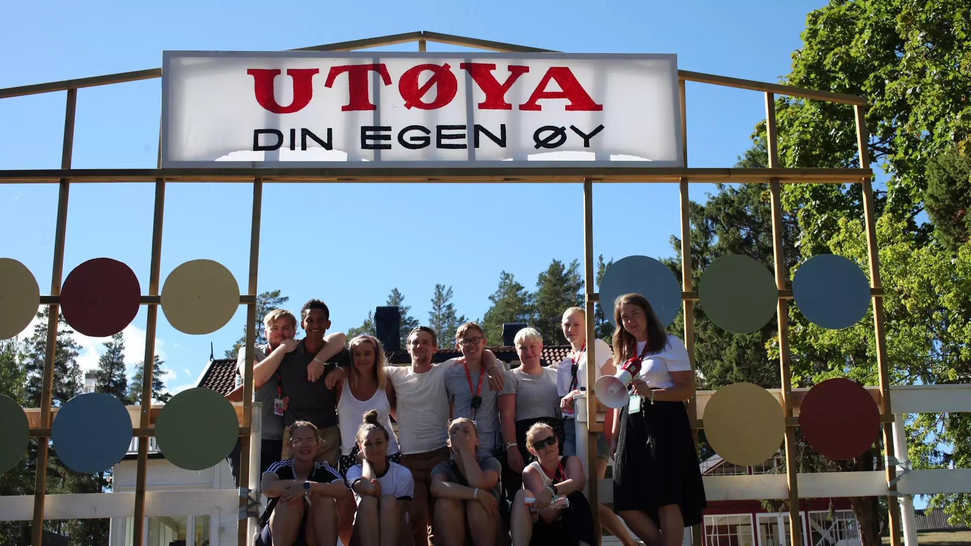 En gjeng ungdommer poserer ved en port som har et skilt på toppen der det står "Utøya - din egen øy". Det er fint vær og sommer. 