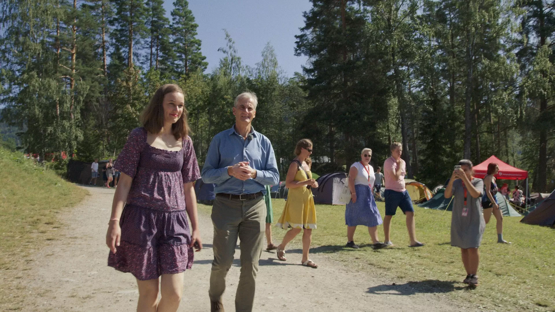 Jonas Gahr Støre og Ina Libak går langs en grusvei på Utøya en sommerdag. Bak dem ser man ungdom og telt. 