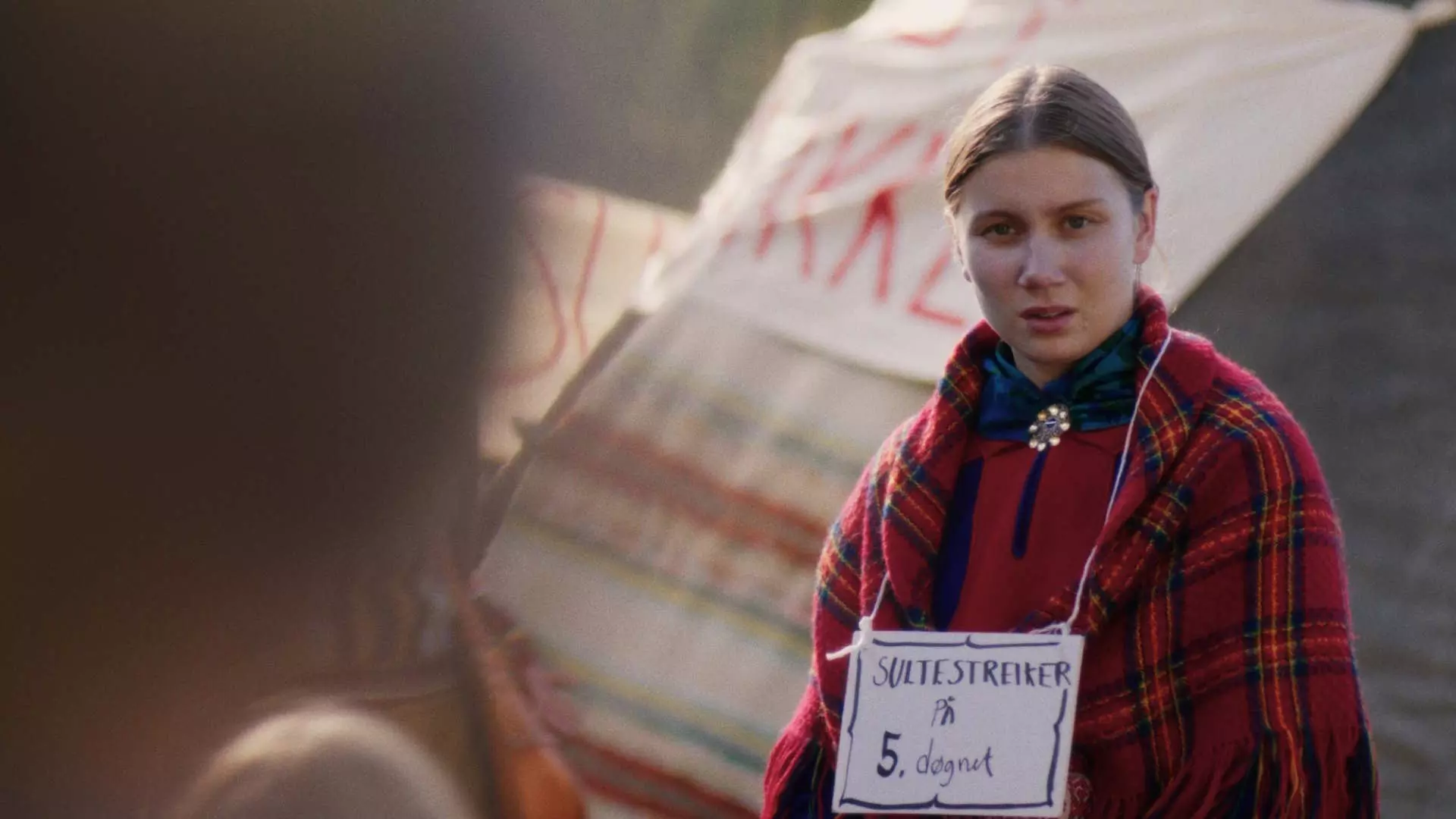 Ung kvinne ikledd rødt rutete sjal og en samisk sølje i halsen. Rundt halsen henger et skilt hvor det står "sultestreiker". Hun ser oppgitt ut. I bakgrunnen kan vi skimte en lavvo. 