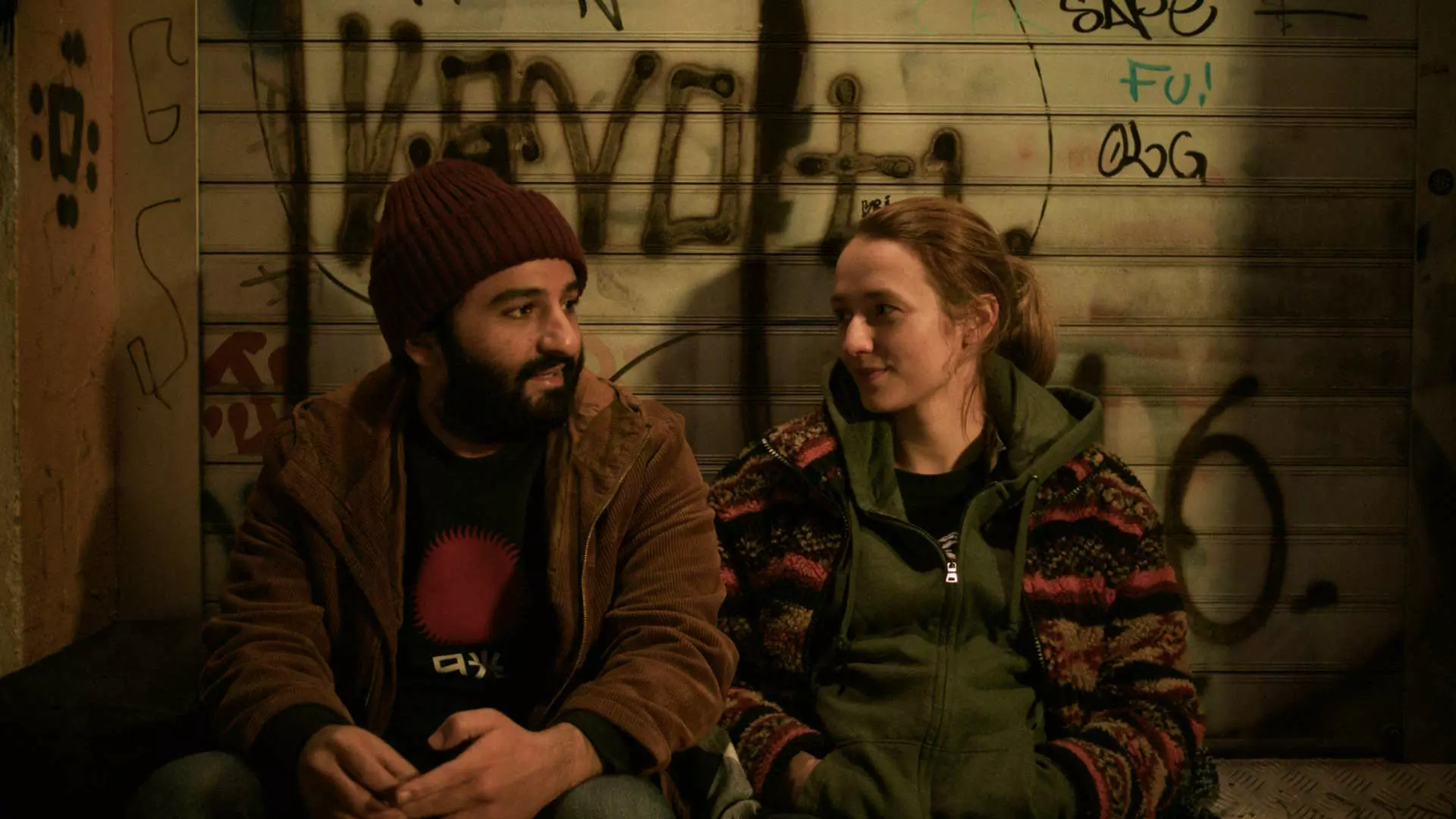 En ung mann og kvinne sitter på en benk. Veggen bak dem er rekket i grafitti. De ser ut til å ha en hyggelig samtale sammen.