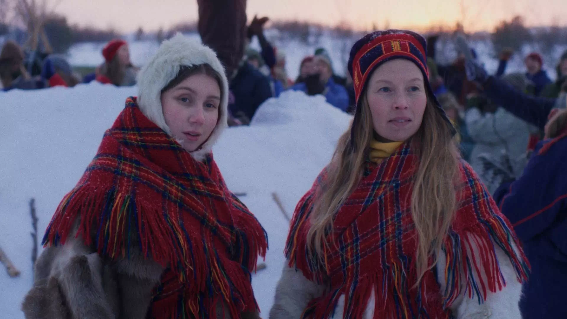 To unge samiske kvinner. De befinner seg ute. Det er snø. De har på seg røde rutete sjal og pelskåper.