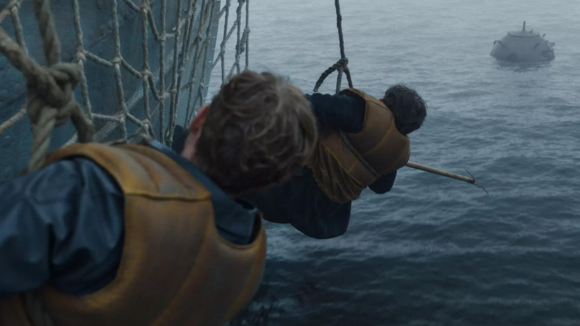To menn henger i et nett på utsiden av et stort skip med ryggen til kameraet. Den ene mannen har en lang pinne med en krok på. Det ser ut som de prøver å hente noe eller noen som vi ikke ser opp fra havet.  
