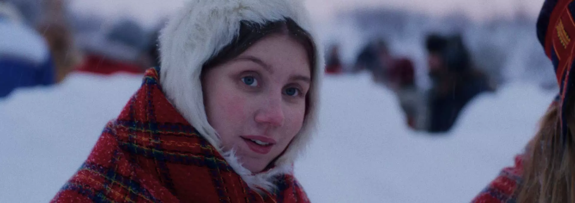 Ung samisk kvinne. Hun har en hvit pelslue og et rødt rutete sjal på seg. Hun er ute. Det er snø rundt henne.