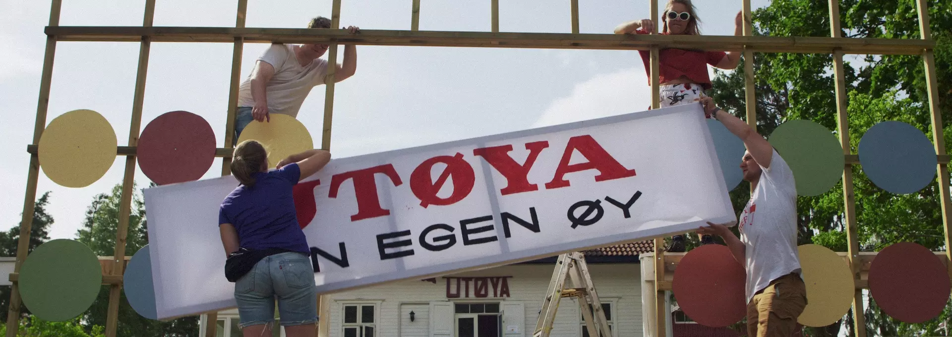 Fire ungdommer står og prøver å henge opp et skilt som det står "Utøya - din egen øy" på en slags port. Bak dem kan du se det hvite huset som er på Utøya. 