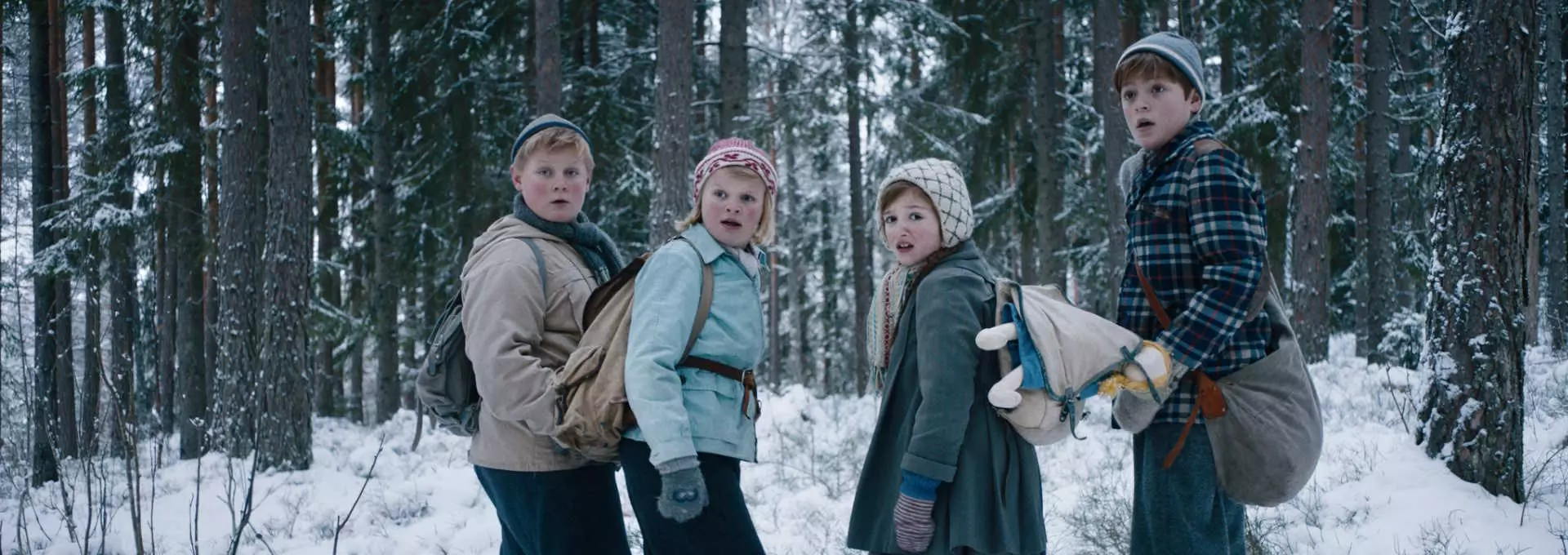 Halvtotal av fire barn som står i en snødekket skog. De er ikledd vinterklær fra gamledager. De snur seg over skulderen og ser sjokkerte ut. Foto.