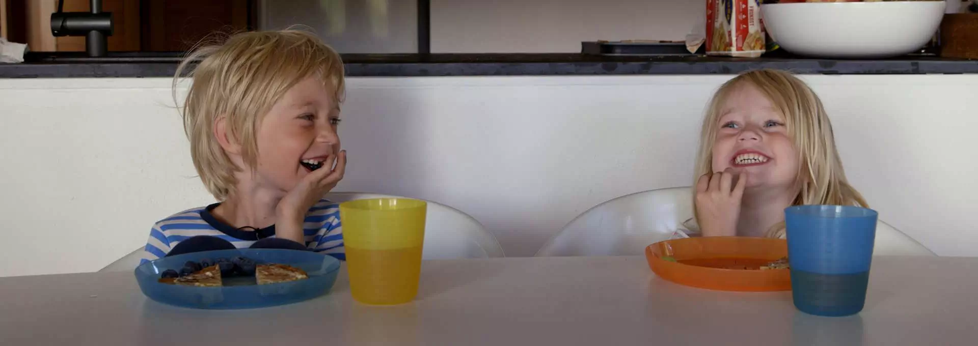 To barn sitter ved frokostbordet. De ler og koser seg. Forran dem på bordet har de hver sin plastkopp og plastikktallerken. Foto
