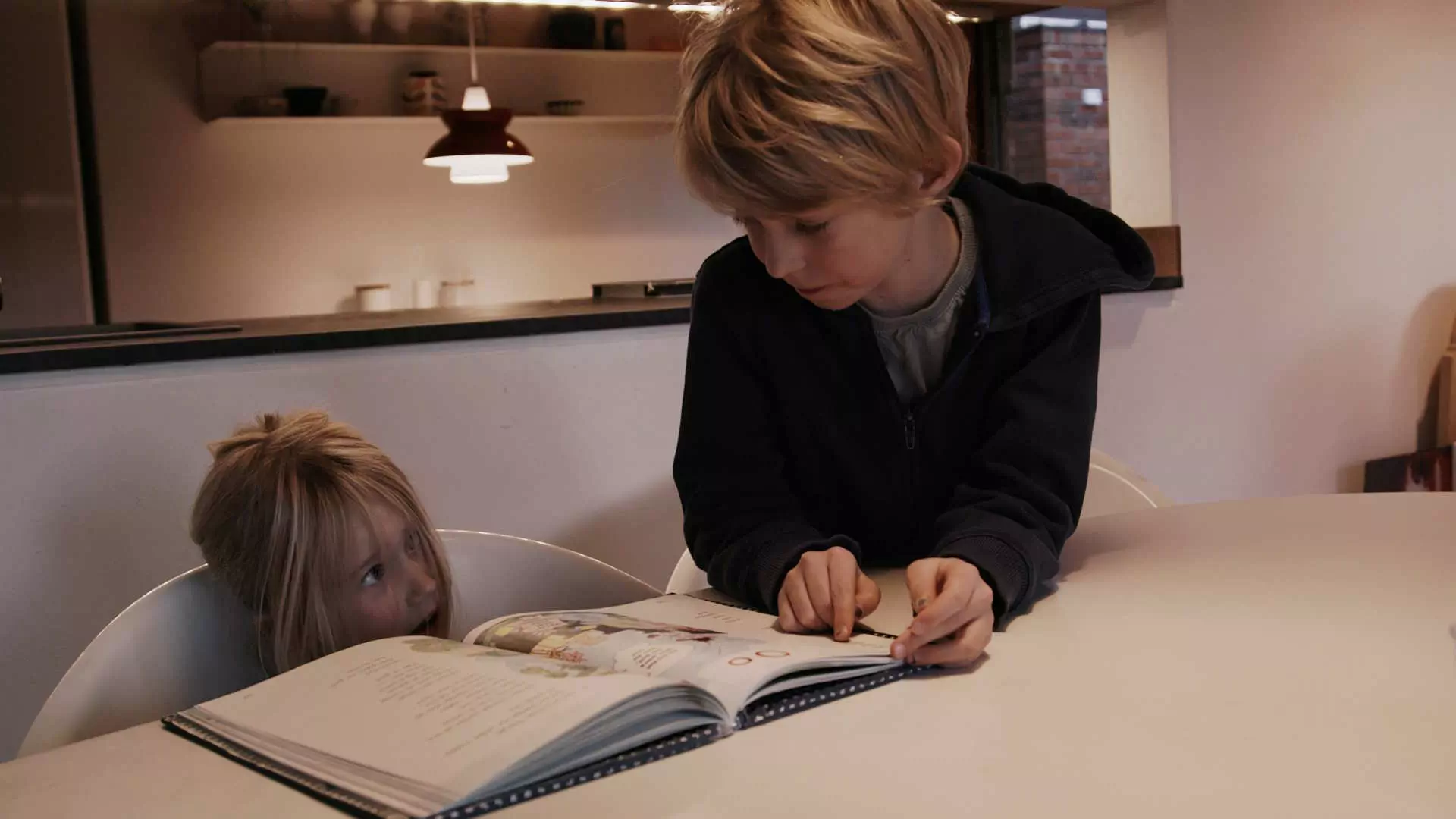 To barn sitter ved et spisebord. På bordet ligger en åpen bok. Gutten peker i boken som om han prøver å forklare noe til jenta. De ser på hverandre. Foto.