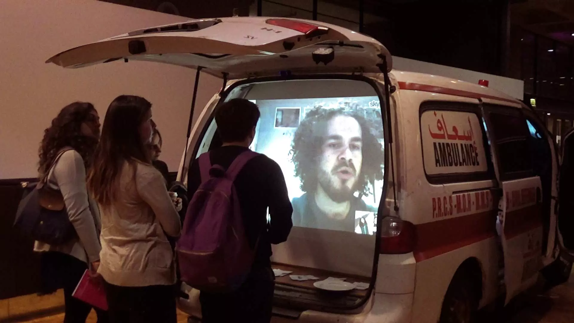 Stillbildet hentet fra en scene i "Livet er vakkert". En tv-skjerm i bagasjerommet av en varebil. Tre mennesker ser på skjermen. Nærbilde av en mann på skjermen. Foto.