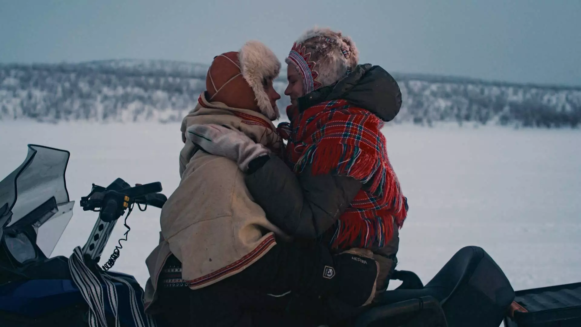 Halvtotal. En samisk mann og samisk kvinne sitter ansikt til ansikt på en snøscooter. De holder rundt hverandre og ser forelsket ut. I bakgrunnen ser vi snø og fjell. De har på seg tradisjonelle samiske plagg. Foto.
